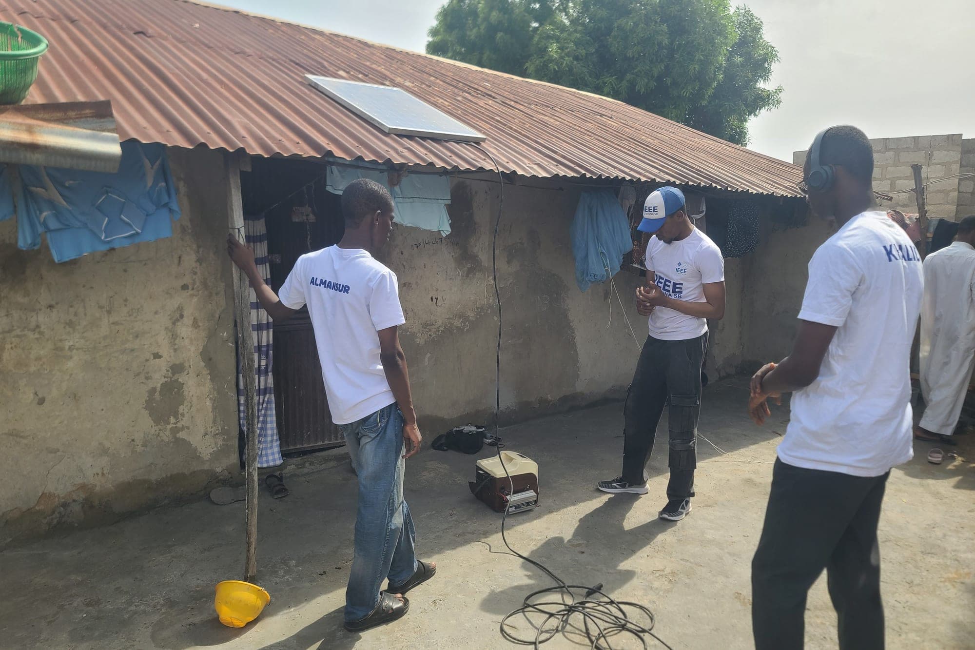 SolarPeer team members check a power unit on the ground, connected by a cable to a solar panel on the roof.